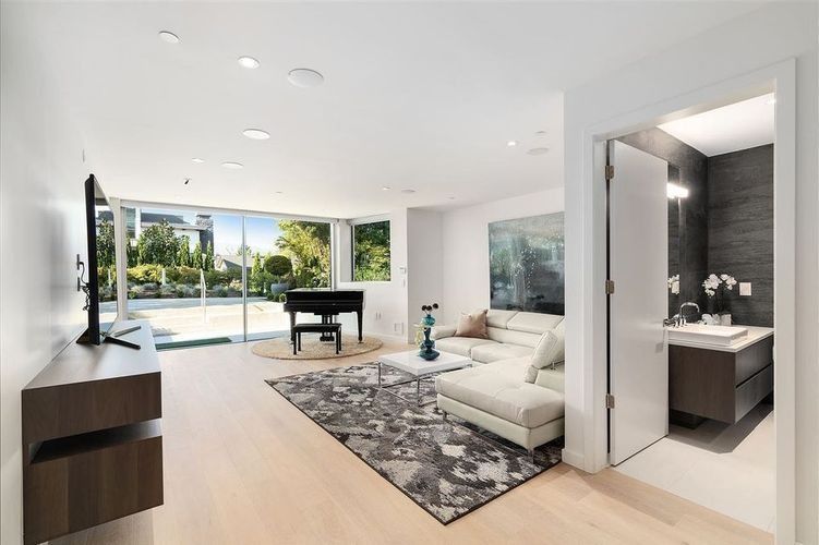 Modern open-plan living room with white furniture, piano, large windows, and outdoor patio view