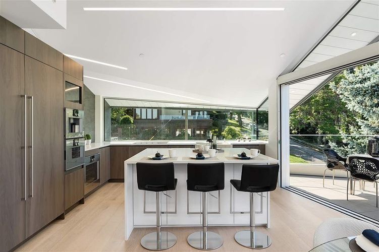 Modern kitchen with island, black stools, wood cabinets, and large windows overlooking greenery