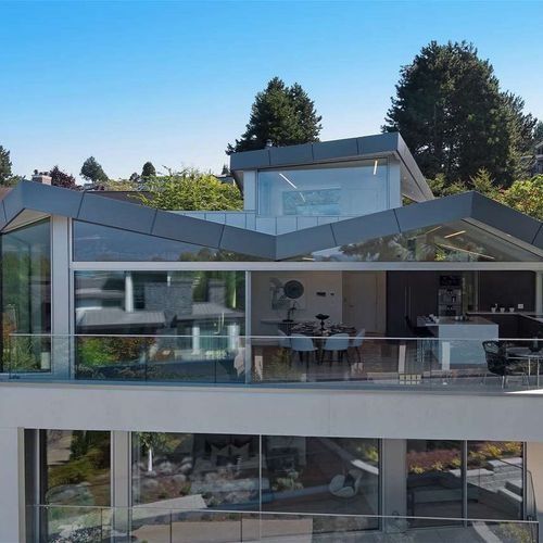 Modern glass house with angular roof and outdoor patio, surrounded by trees under a clear blue sky