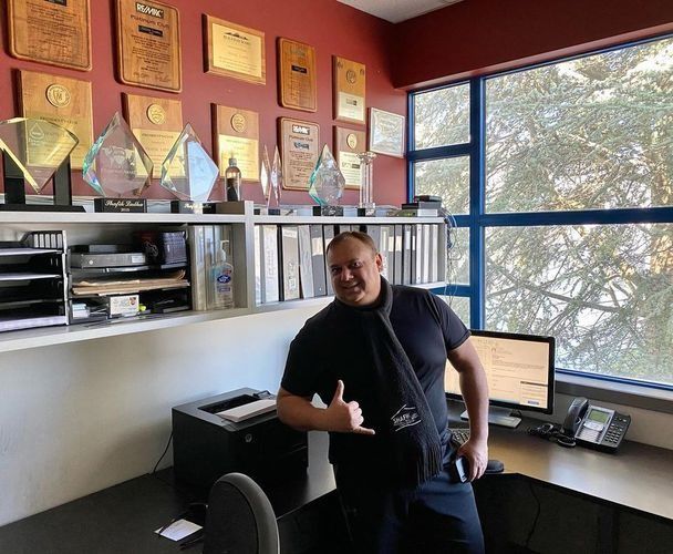 Man standing in a sunlit office beside a desk, with framed awards on the wall and a computer monitor nearby.