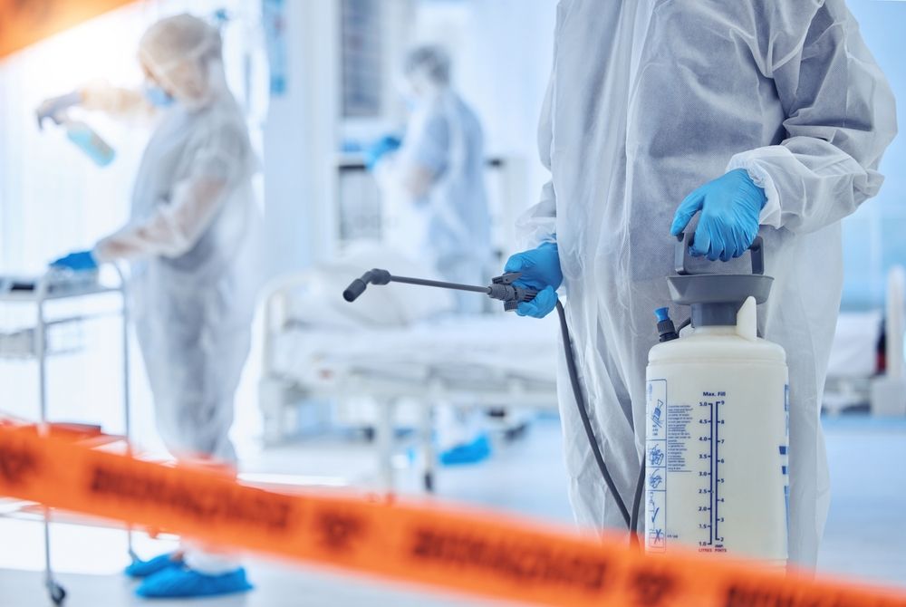 Protective-suited workers spraying disinfectant in a bright lab, with a caution tape barrier in front.