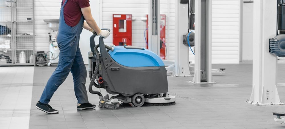 Worker pushing a blue-gray floor scrubber in a bright industrial facility