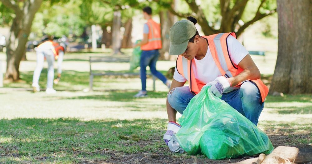 Worker in orange safety vest kneels to collect trash into a green bag in a park.