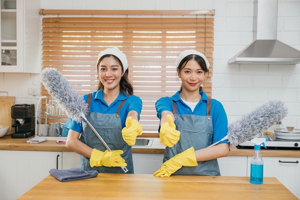 Two cleaners in blue uniforms and yellow gloves tidying a kitchen counter with dusters and spray bottle