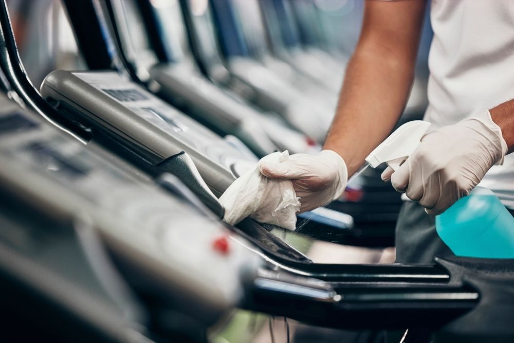 Gloved hands cleaning treadmill consoles in a gym with a blue spray bottle