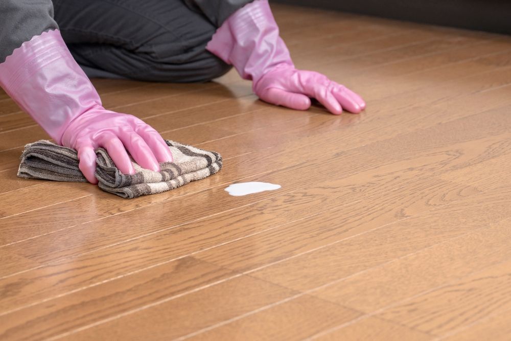 Pink-gloved hands wiping a wooden floor with a cloth.