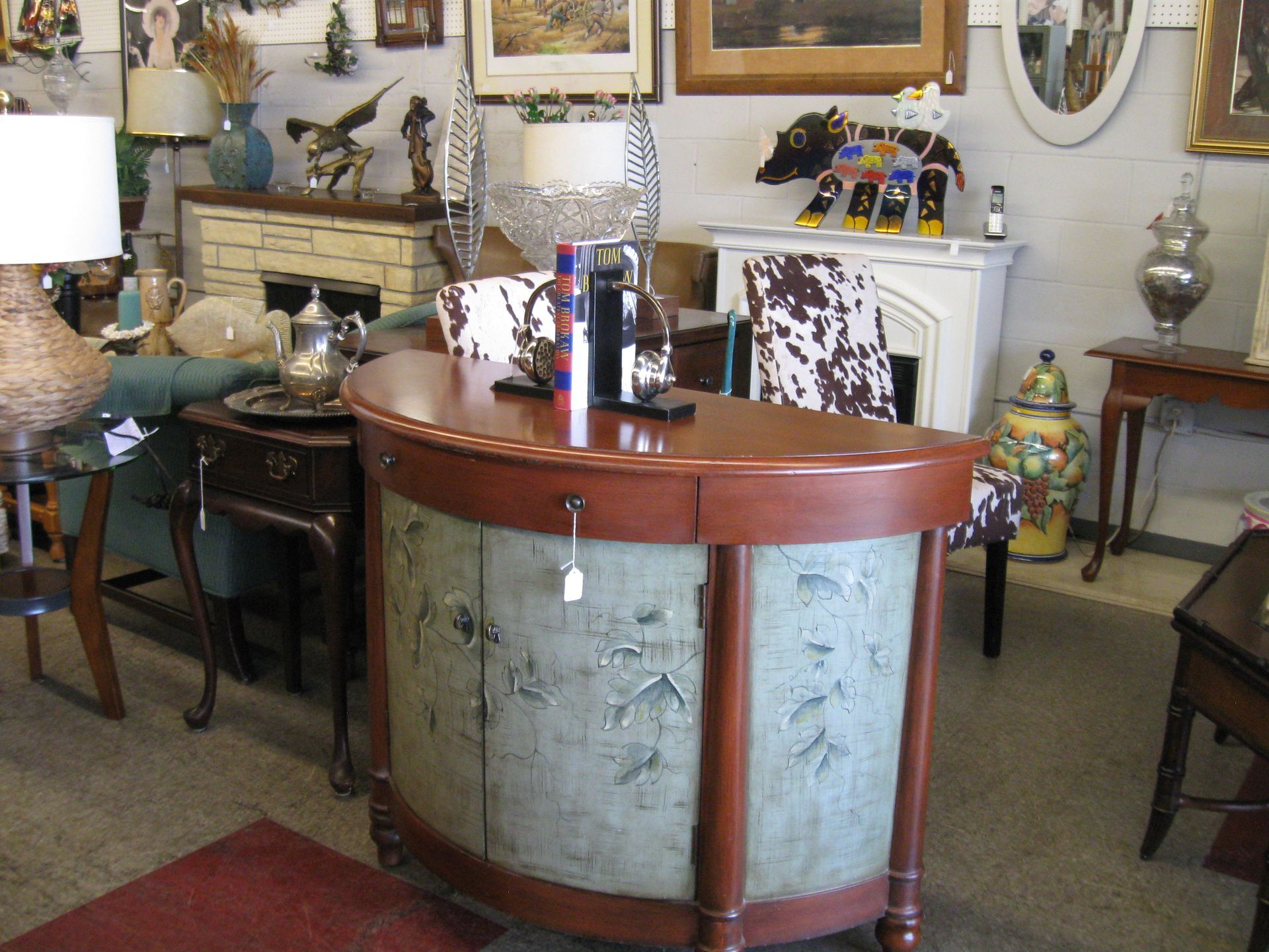 An antique-style table in a cluttered vintage room with lamps, mirrors, and framed art on the walls