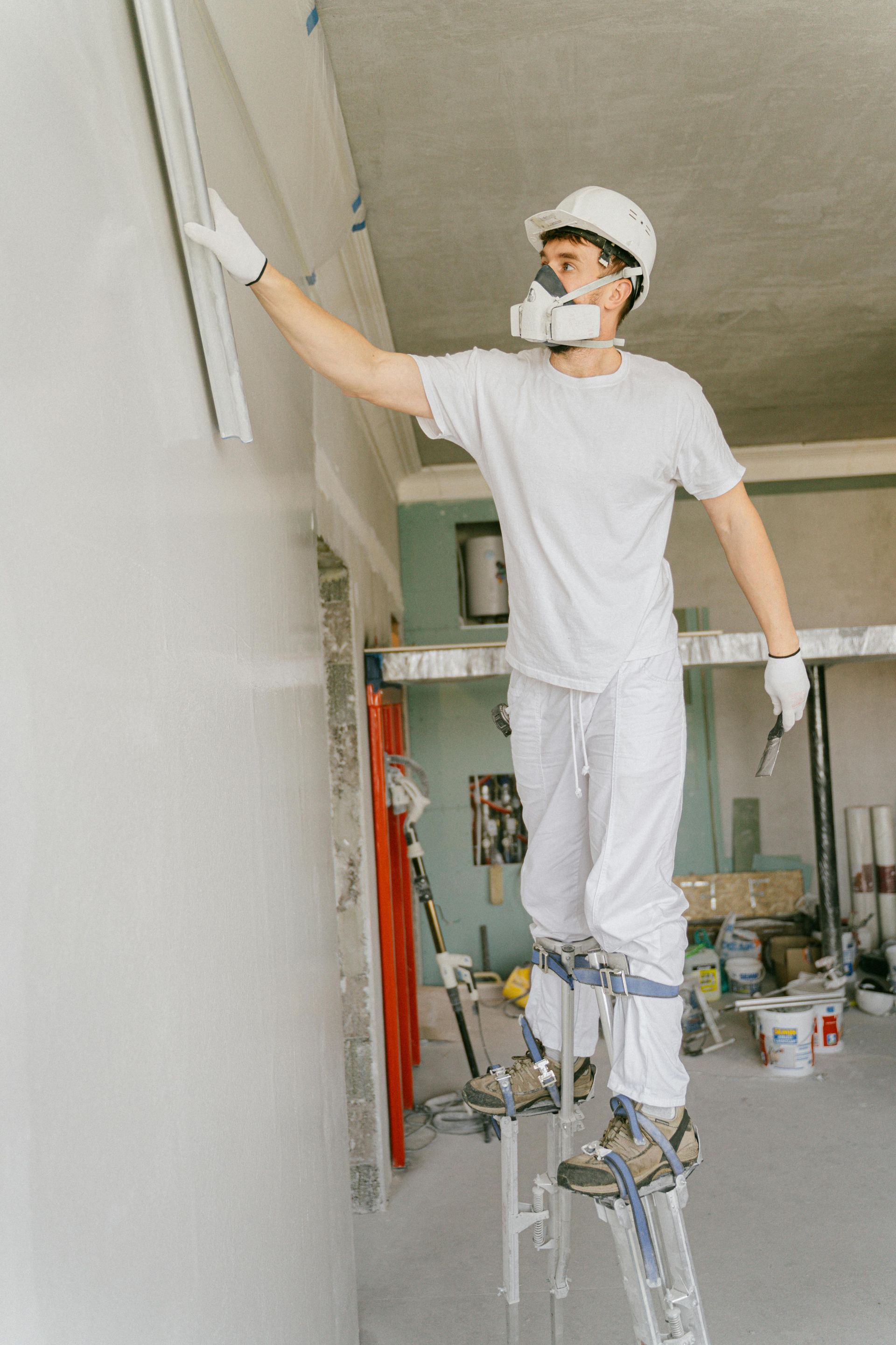 Construction worker on stilts, applying plaster to a wall indoors. Wearing respirator, gloves, and hard hat.