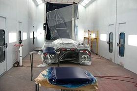 Car under repair in a spray booth, with a hood open and covered parts in the foreground.
