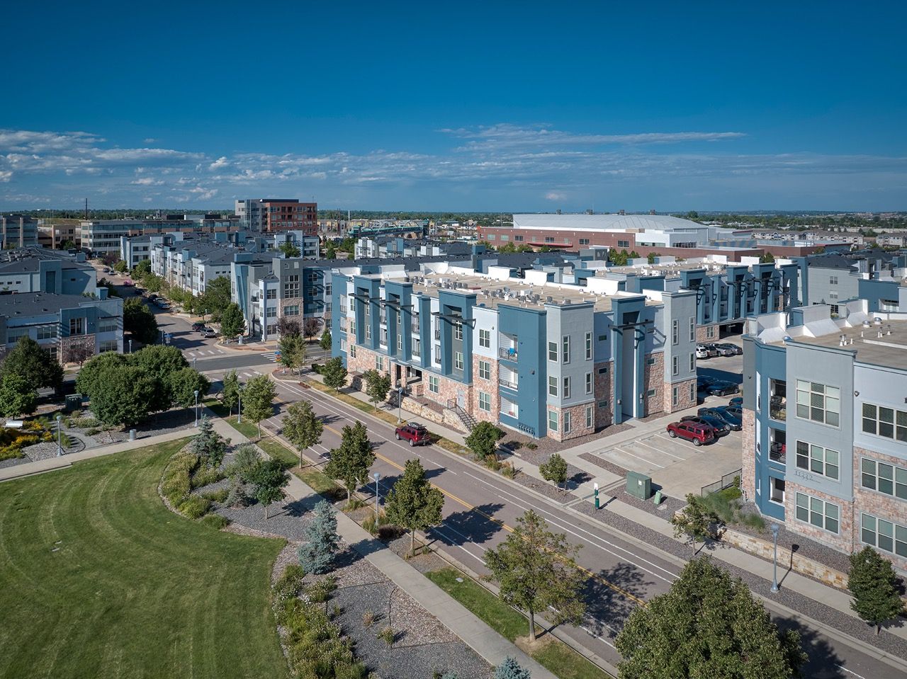 Aerial view of a modern apartment community with blue-and-stone buildings, landscaped spaces, and tree-lined streets.