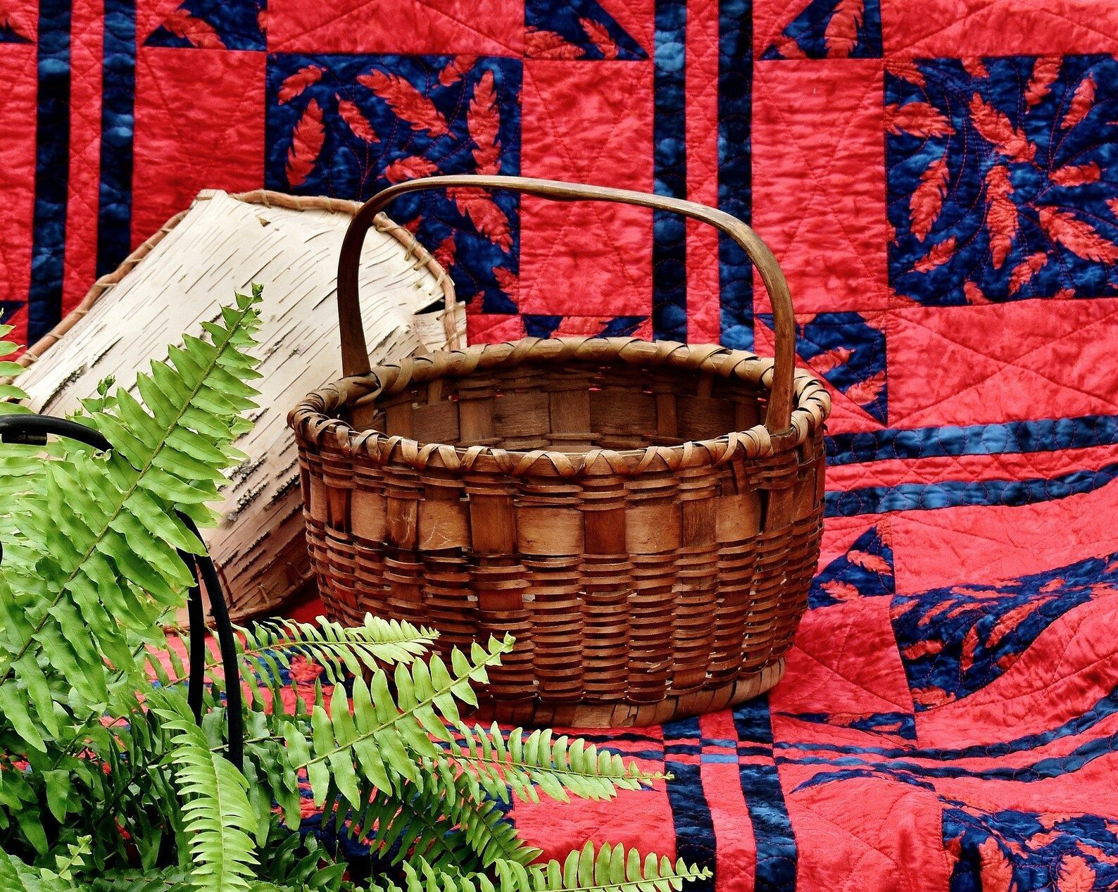 Woven basket and folded white cloth on a red patterned quilt with green fern leaves