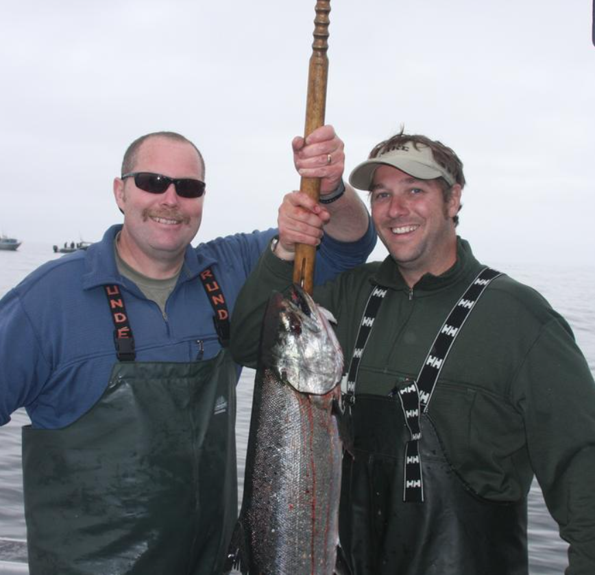 Two men in fishing gear hold up a large fish on a boat in snowy conditions