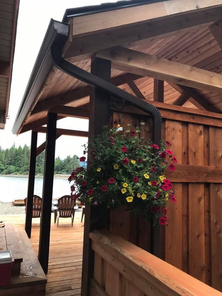 Covered wooden porch with black gutter, hanging flower basket, and lake view beyond