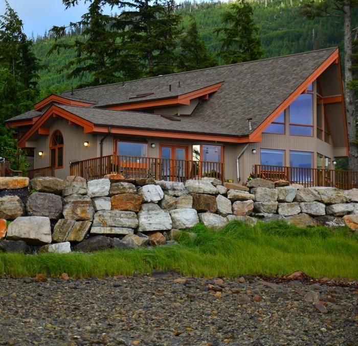 Rustic lodge-style house with orange trim behind a stone retaining wall and green trees