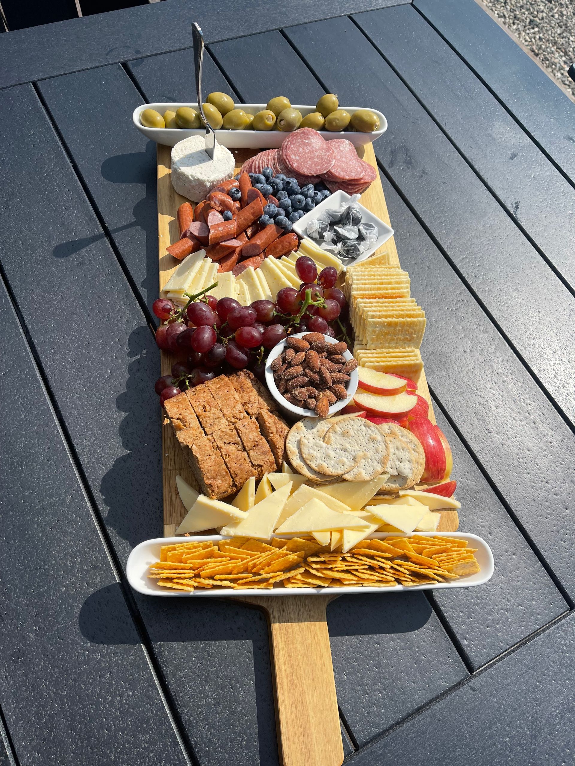 Charcuterie board on a wooden paddle, with meats, cheeses, crackers, grapes, and olives on a dark outdoor table.