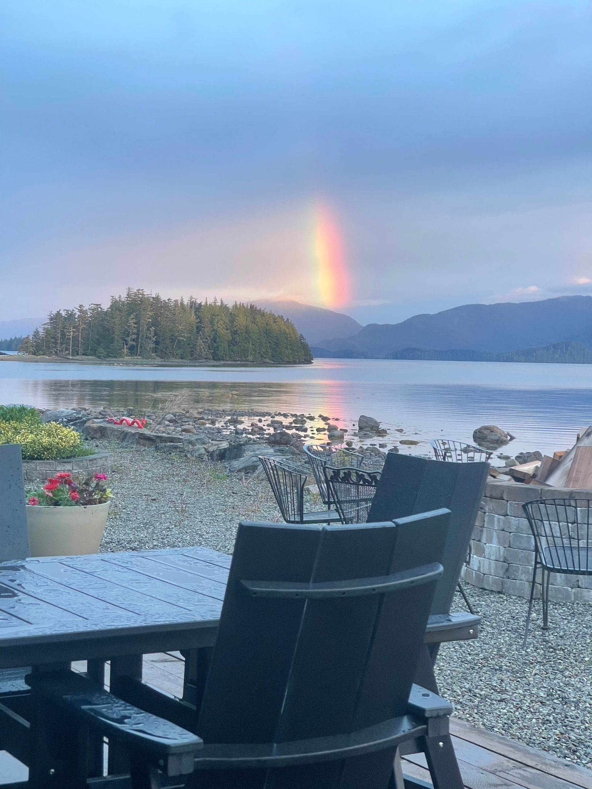 Patio chairs overlooking a calm lake at sunset, with a rainbow over the water and distant mountains