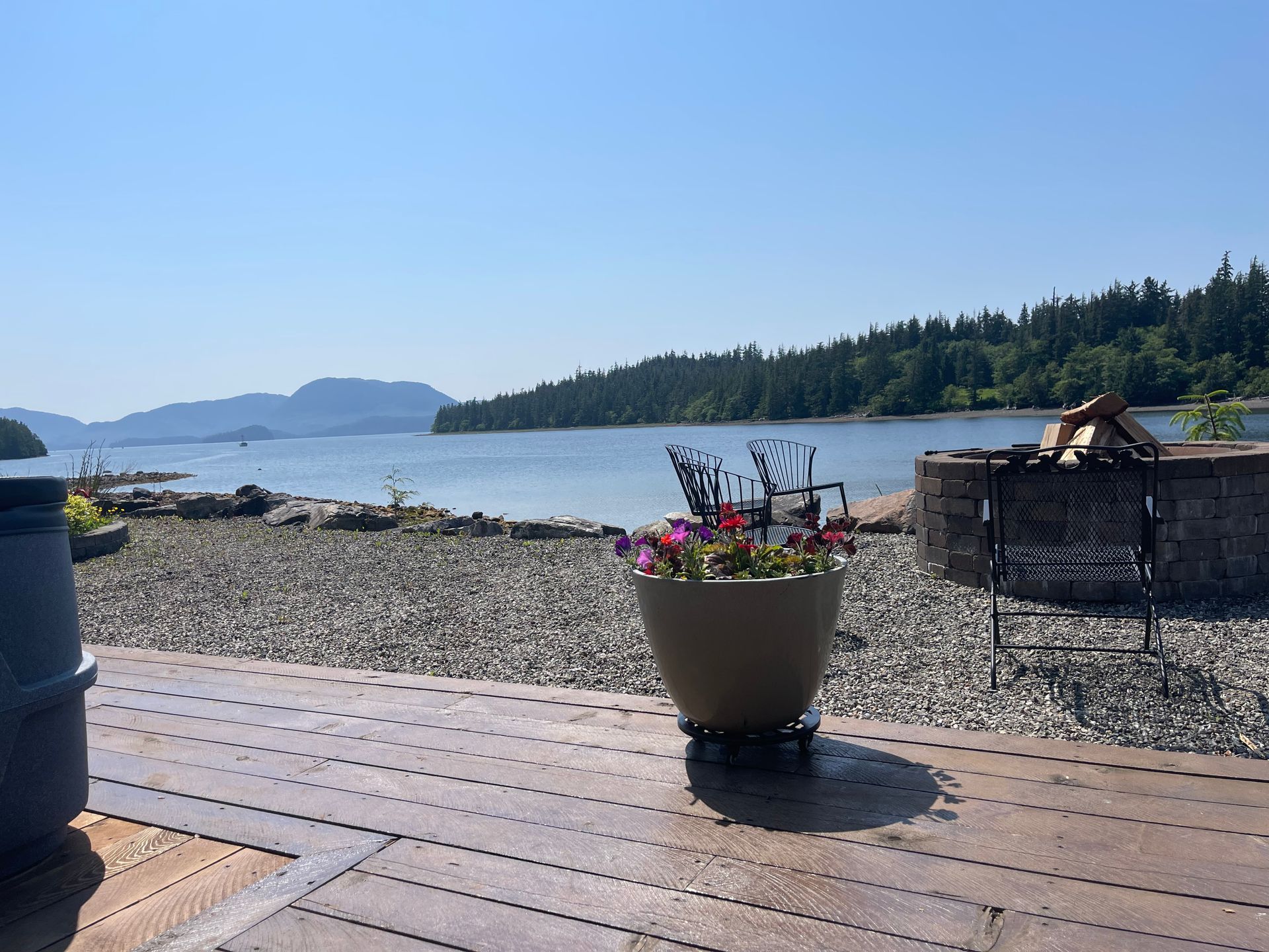 Wooden deck overlooking a rocky shoreline and calm bay, with a potted flower planter in the foreground.