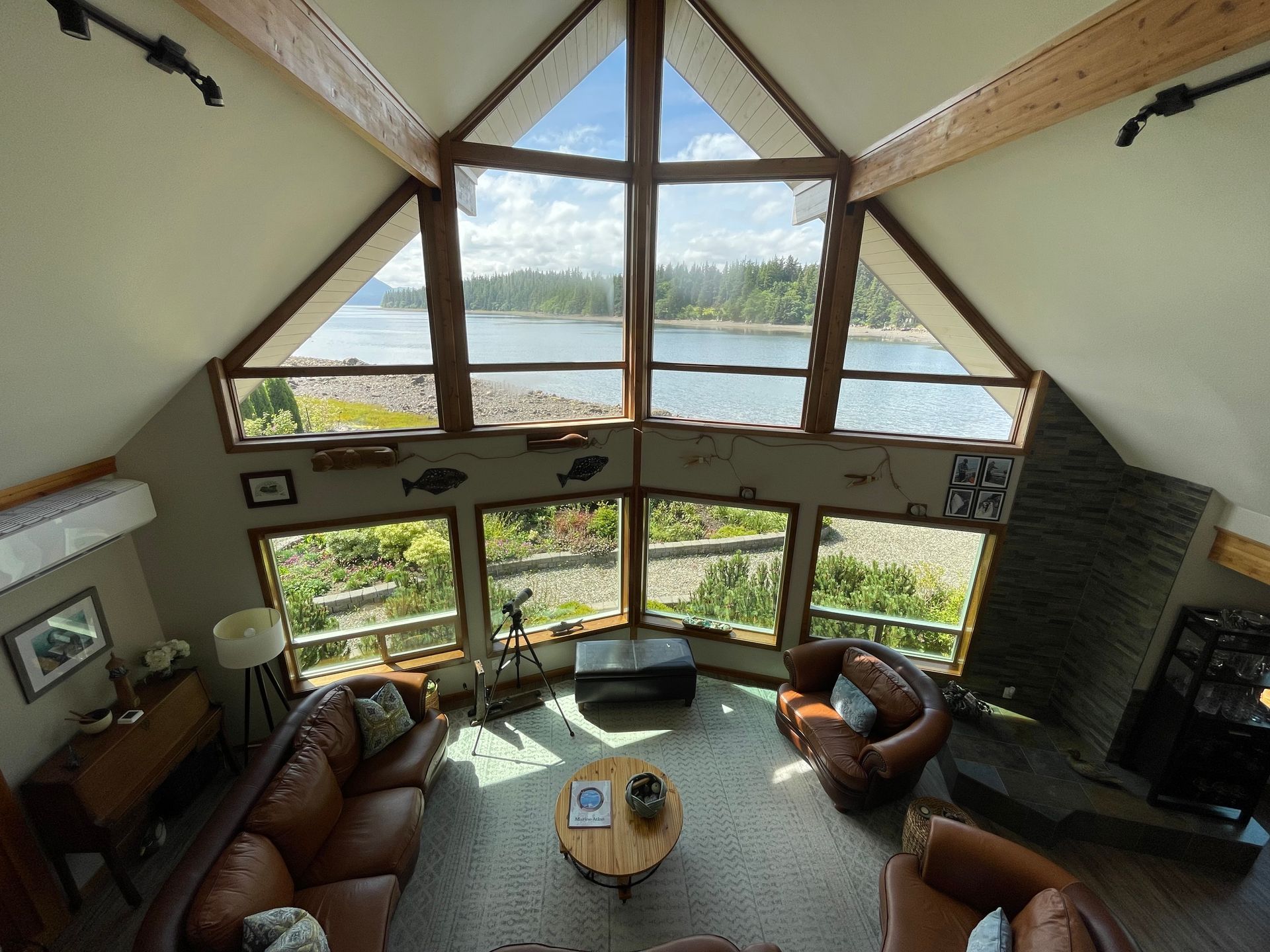A cozy cabin living room with tall windows, brown sofas, a round table, and a lake view.