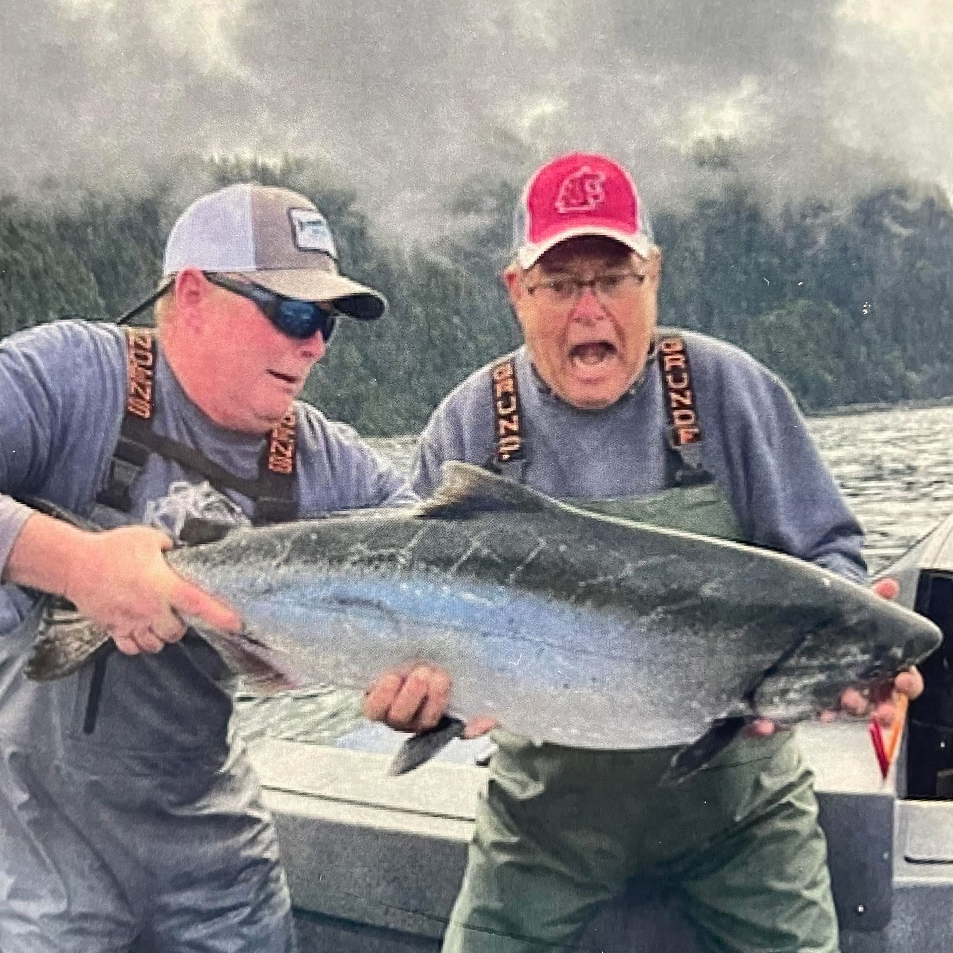 Two anglers holding a large silver fish on a boat, with misty water and trees in the background.