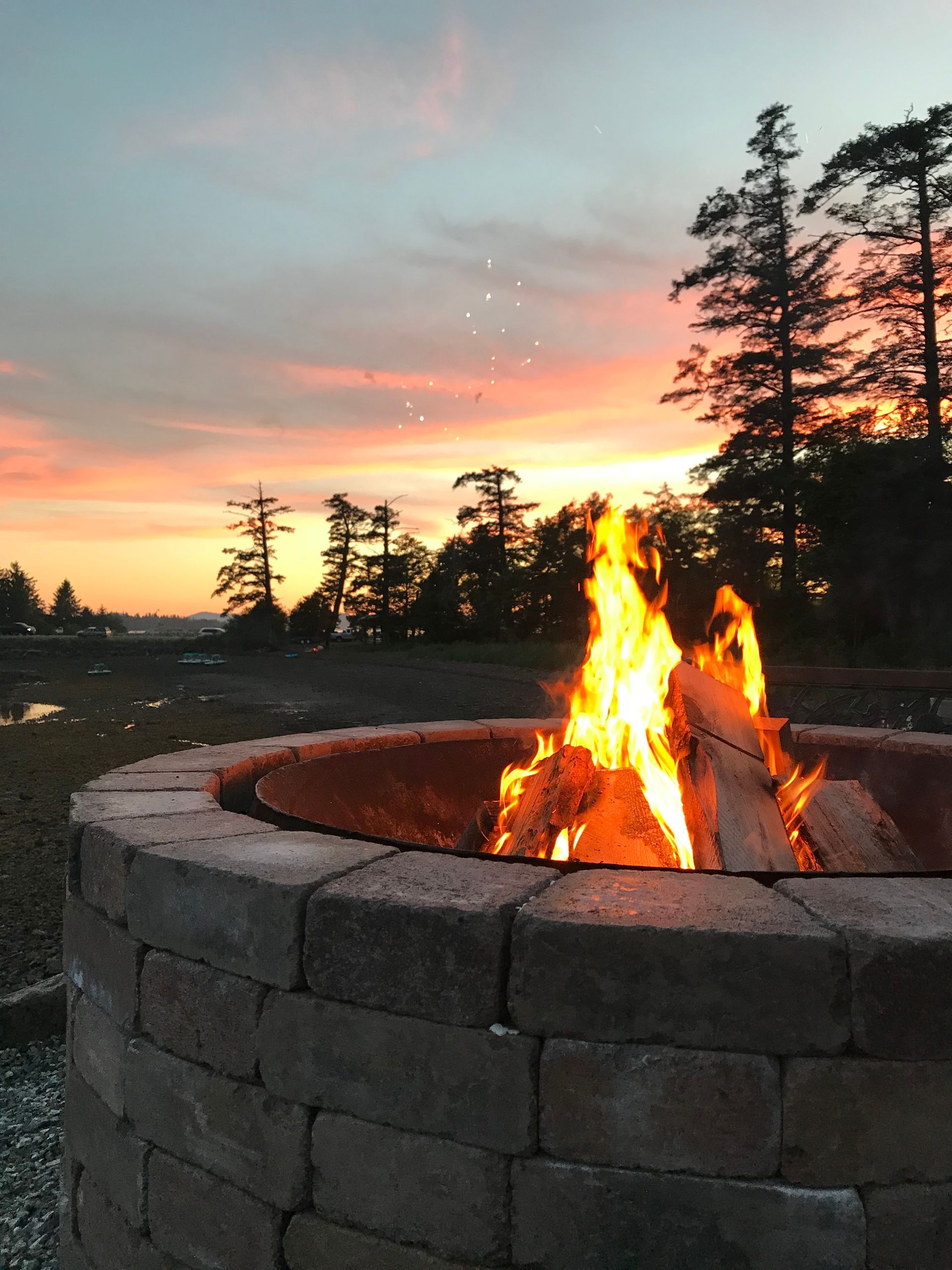 Stone fire pit with a bright campfire at sunset, silhouetted trees and colorful sky in the background