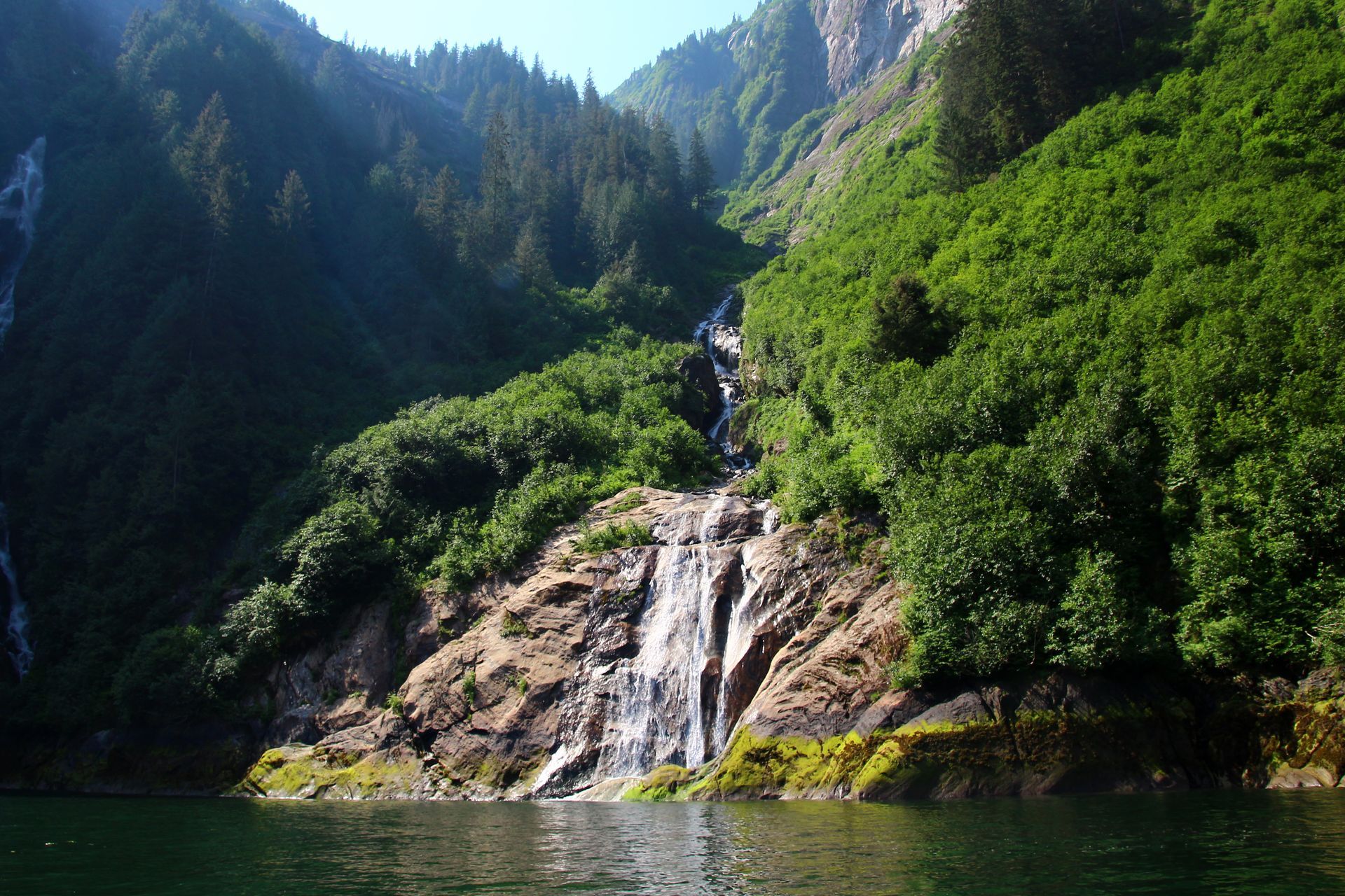 Waterfall cascading over rocky cliffs into a calm lake, surrounded by lush green forest and mountains