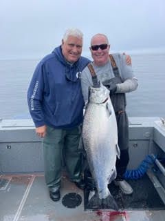 Two men on a boat hold a large silver fish against a gray ocean backdrop