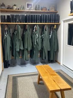 Small mudroom with green coats, wooden bench, and shelves above a row of boots.