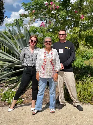 Three people standing outside. Woman in center, two on sides. Plants and flowers in background.