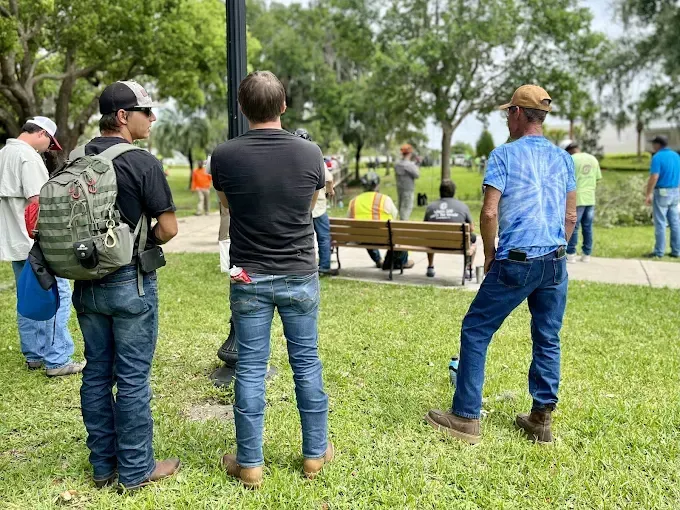 Men standing and conversing outdoors in a park; some wearing work attire. Green grass and trees visible.