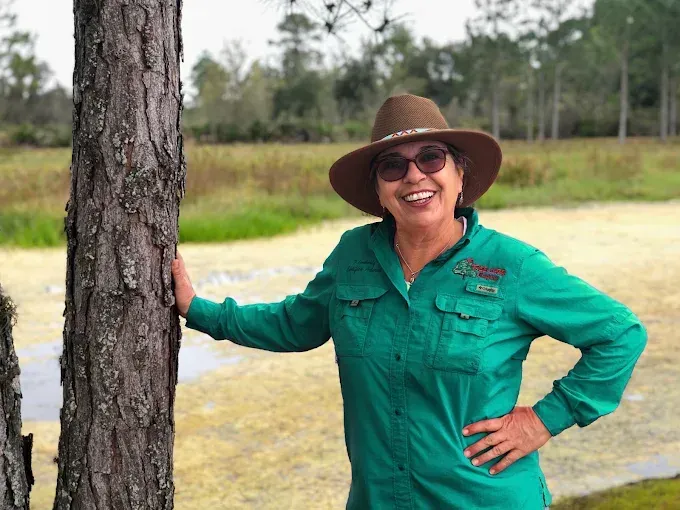 Woman in green shirt and hat, smiling, leaning on a tree trunk outdoors.