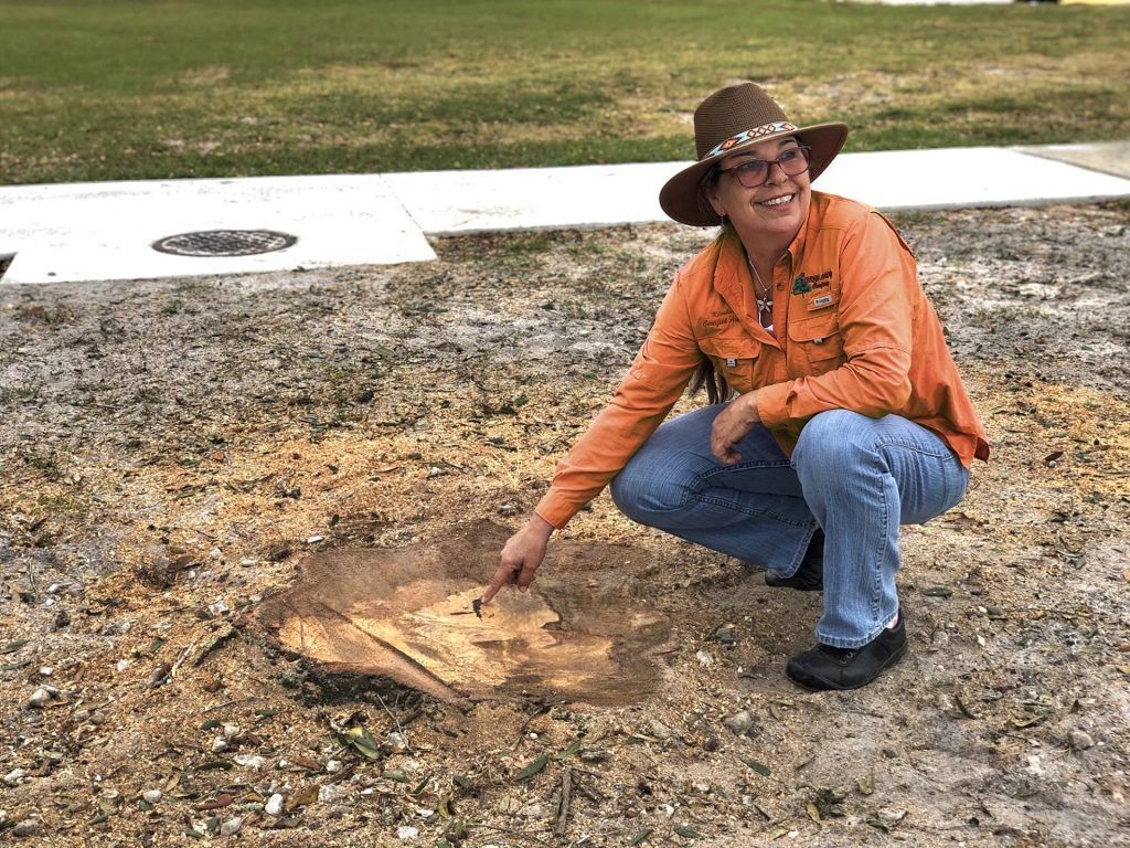 Woman pointing at a hole in the ground; outdoors, wearing hat, orange shirt, and jeans, smiling.