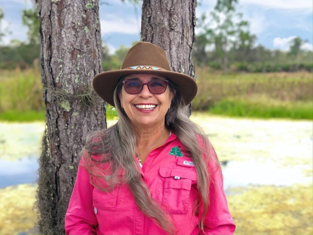 Woman in a brown hat and pink shirt smiles in a swampy area, leaning against a tree.