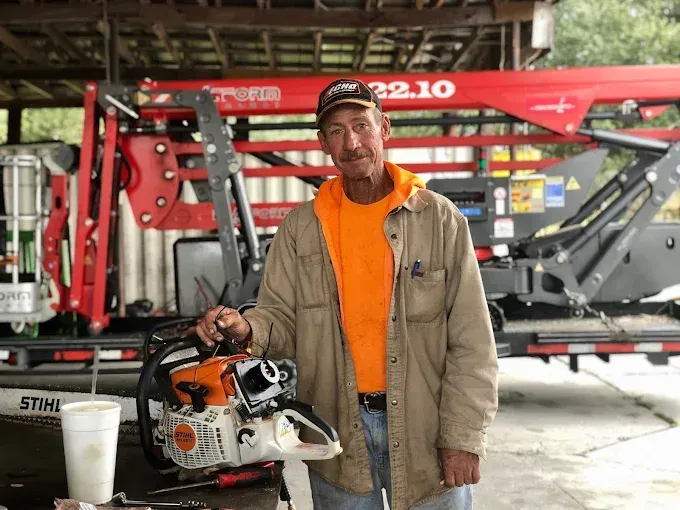 Man holding a chainsaw, wearing orange shirt and tan jacket, standing in front of a red machine.
