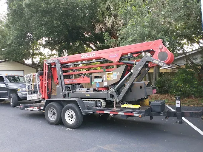 Red aerial lift on a trailer, parked on a street near a house and a truck.