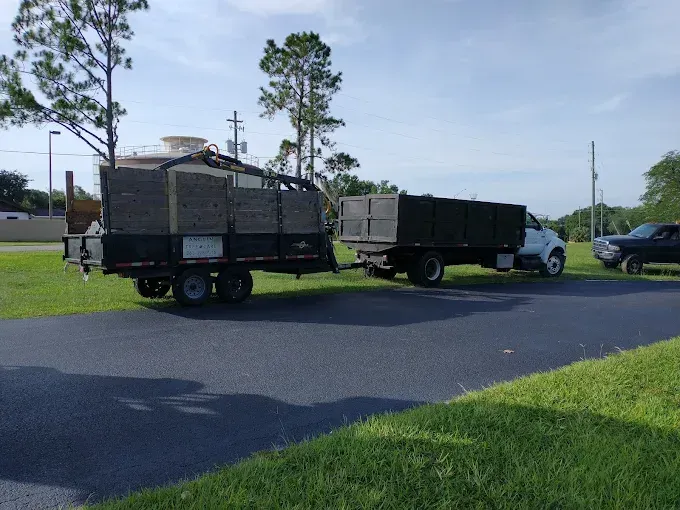 Truck towing a trailer filled with debris parked on a paved area with green grass under a blue sky.