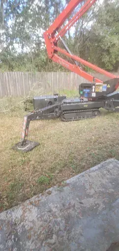 A tracked boom lift on a lawn; fence and trees in background; concrete slab in foreground.