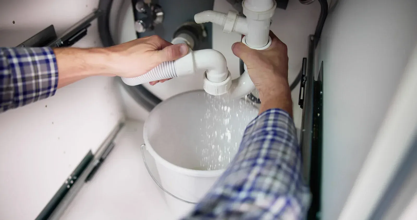 A person in a plaid shirt is unclogging a pipe under a sink, water pouring into a bucket.