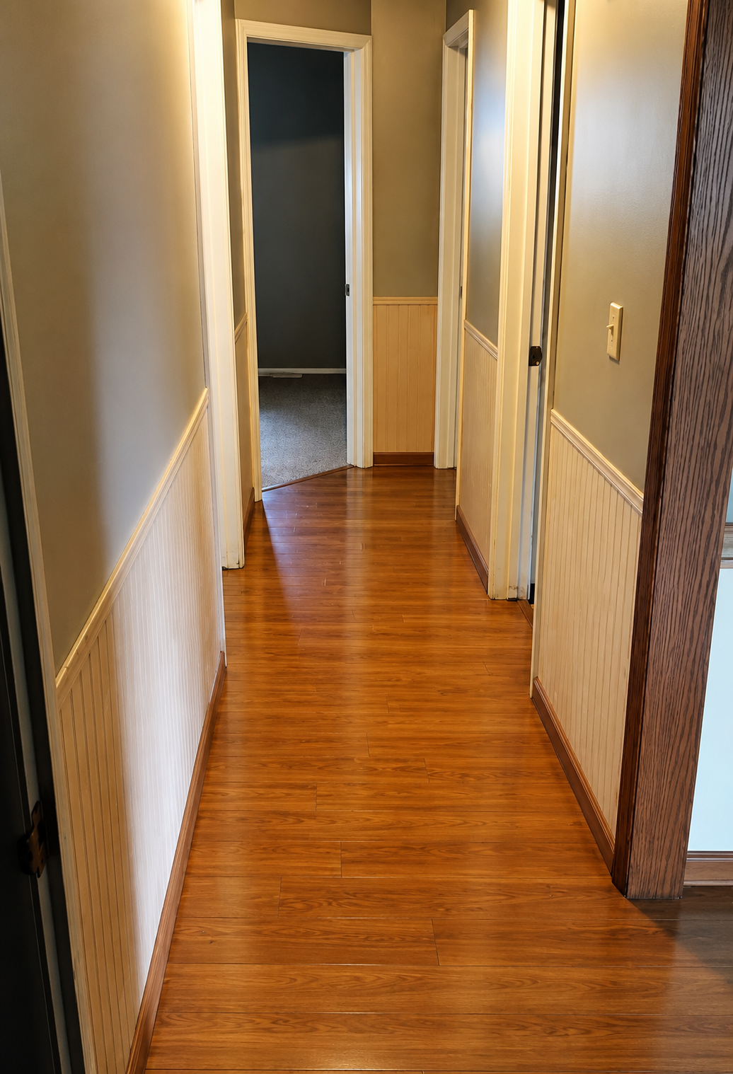 Narrow hallway with glossy wood floors, beige walls, white trim, and open doorways at both ends.