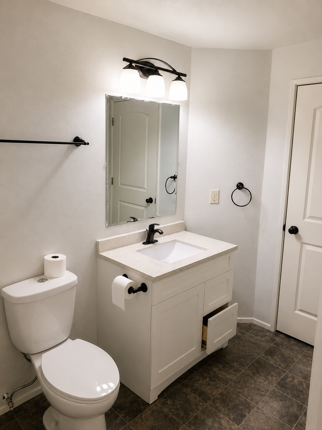 Small white bathroom with vanity, mirror, toilet, towel ring, and dark tile floor.