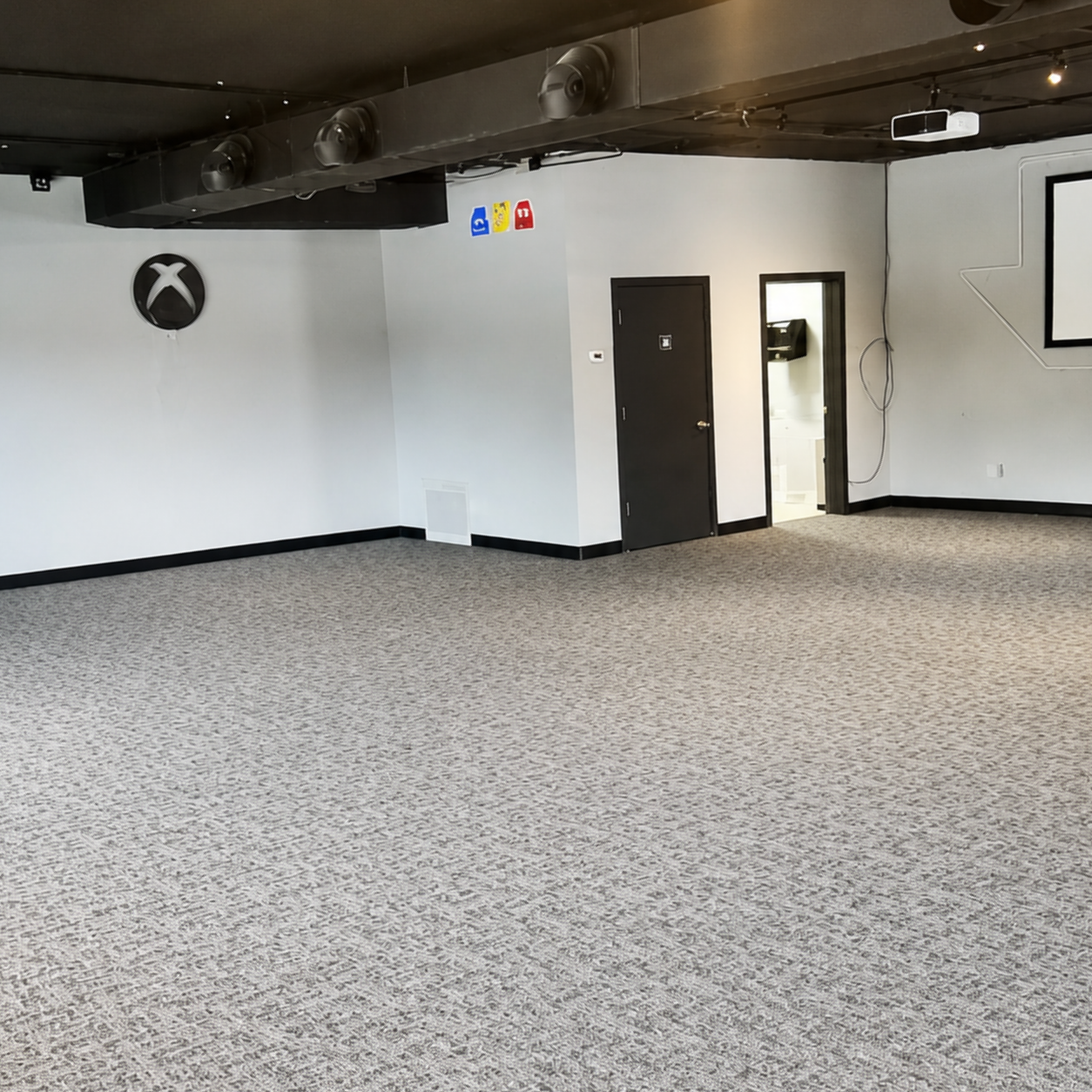 Empty gym room with speckled carpet, white walls, black ceiling beams, and a black door.