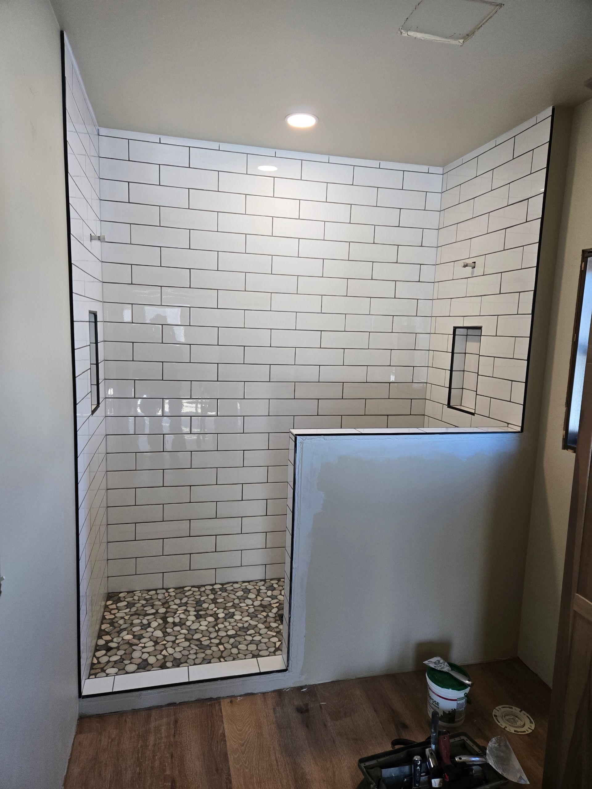 Empty tiled shower under construction with pebble floor, white patterned walls, and half-height partition