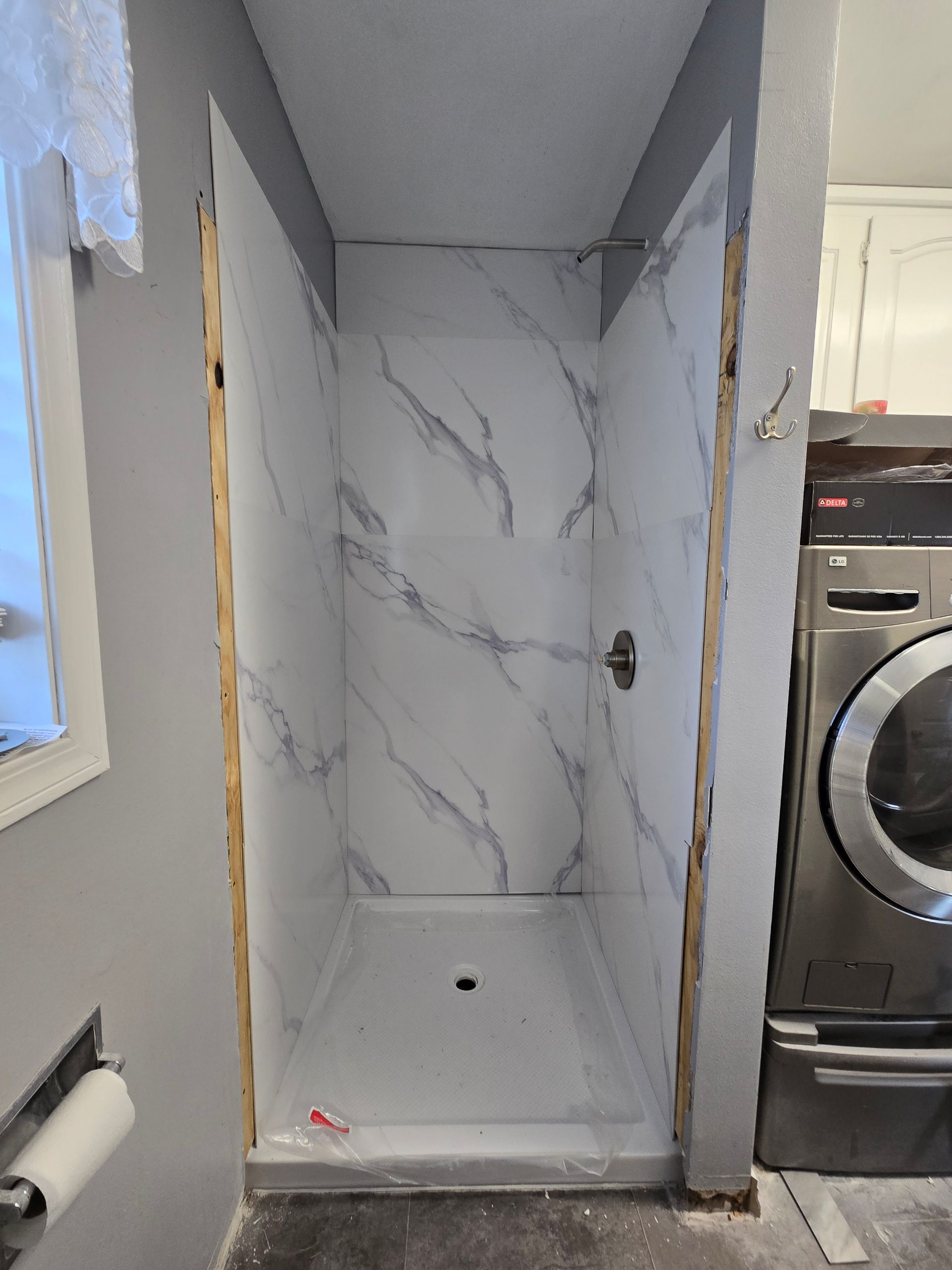 Narrow bathroom shower with marble-look tile, next to a washing machine and gray wall.