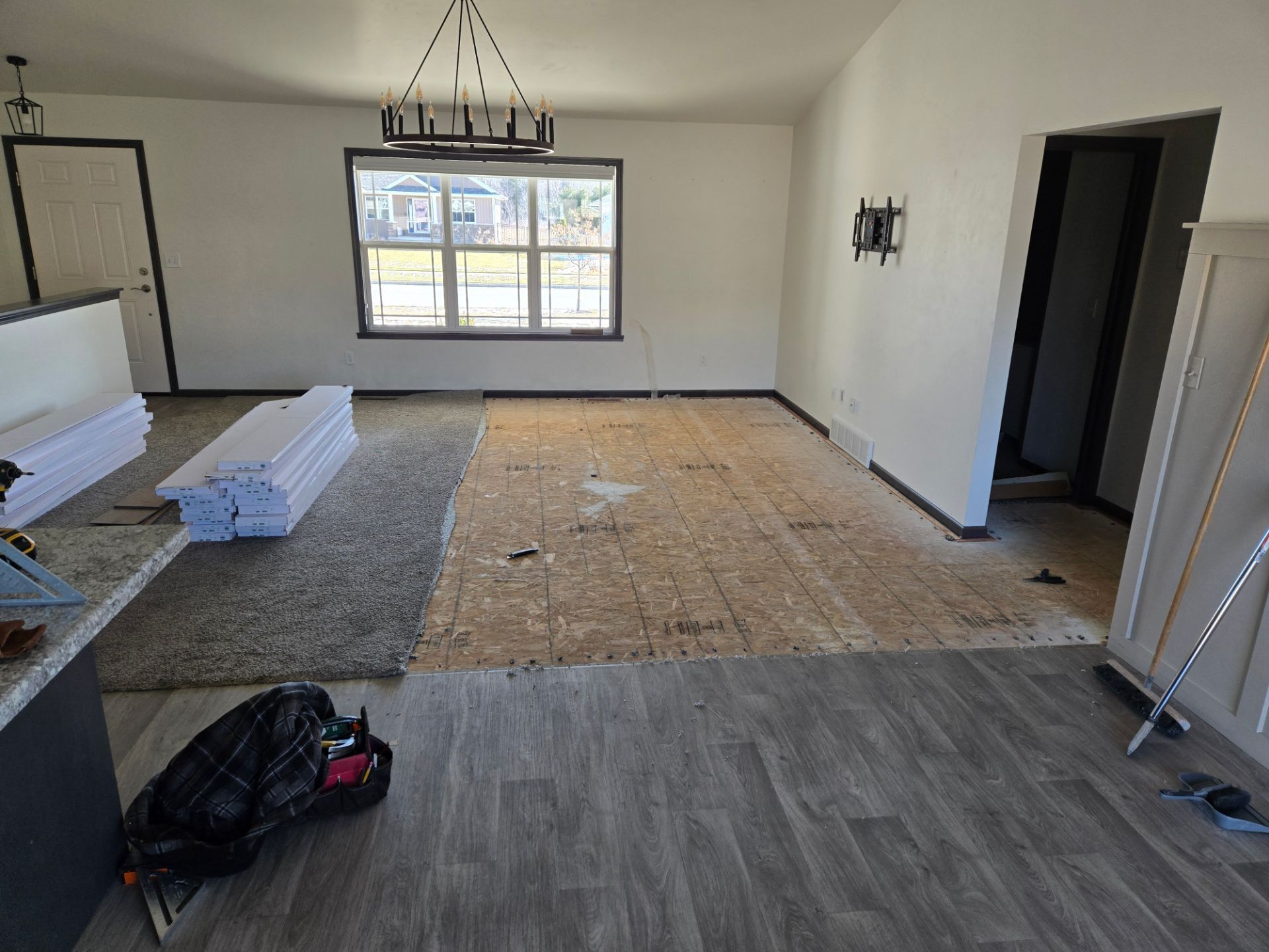Empty living room with gray flooring, exposed subfloor in center, window, and construction materials stacked on the left