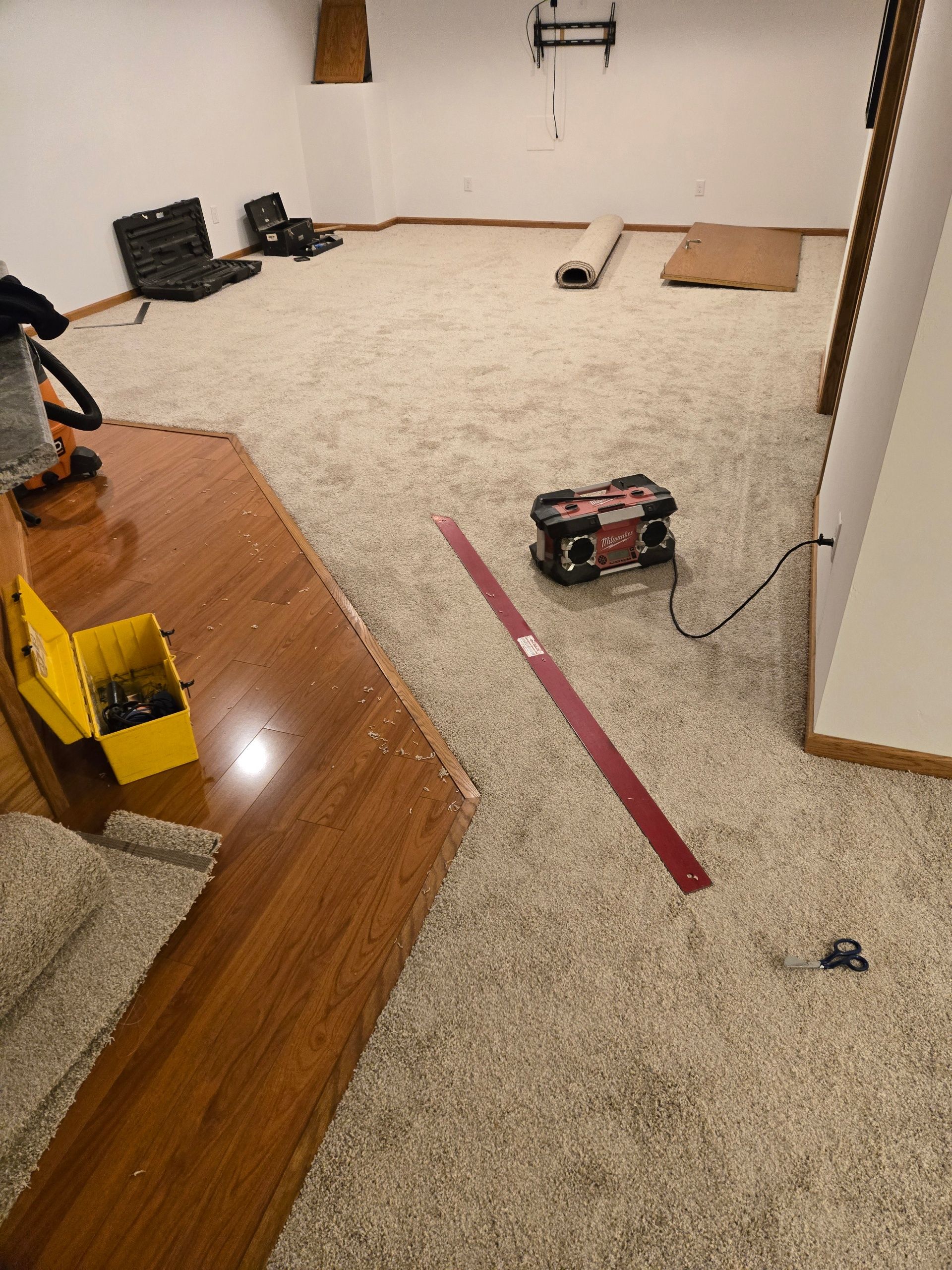 Living room with beige carpet, a red tape line, a toy car, and scattered tools near a hardwood floor.