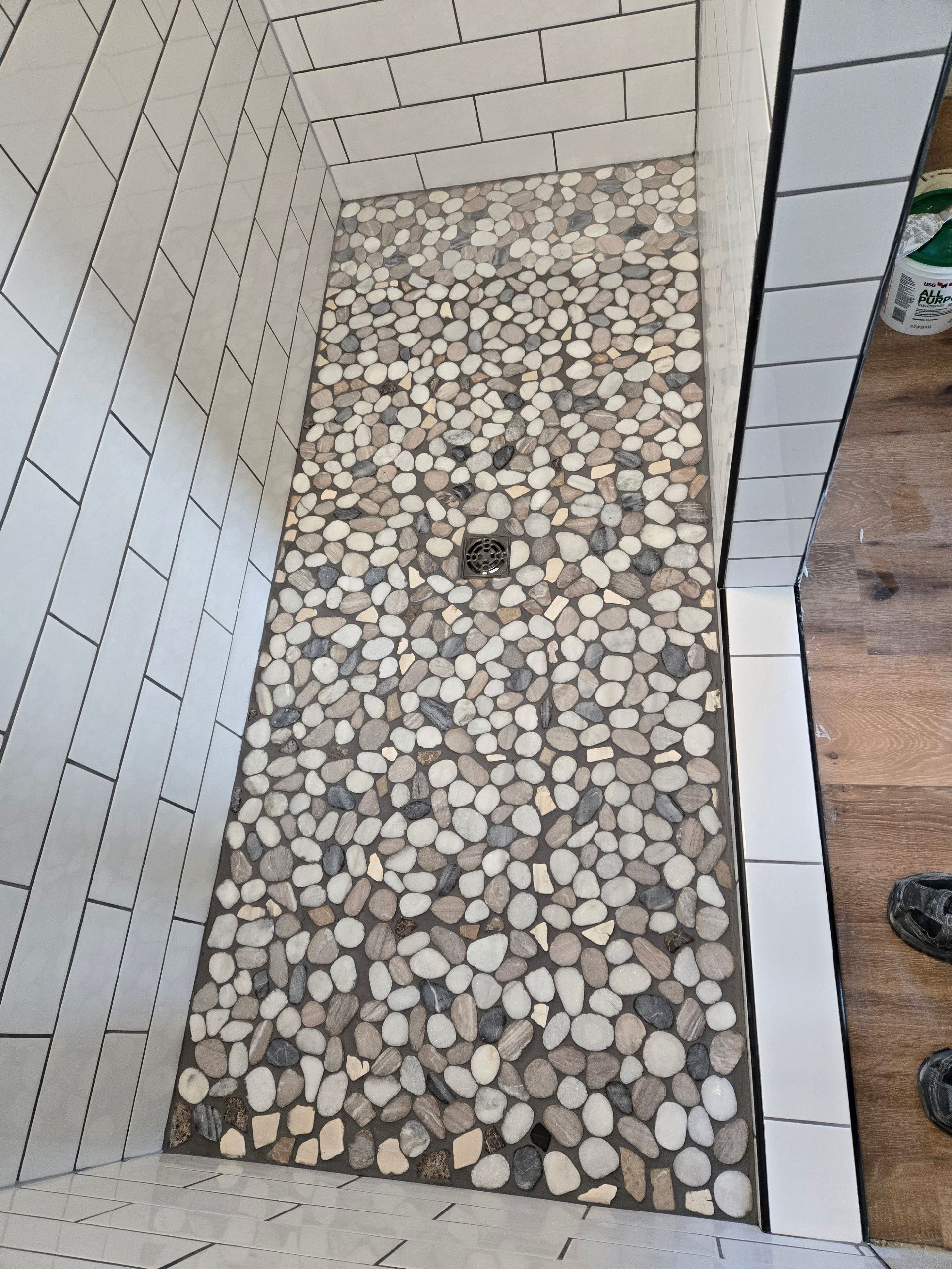 Pebble-lined shower floor with a small square drain, bordered by white tile walls and wood flooring.