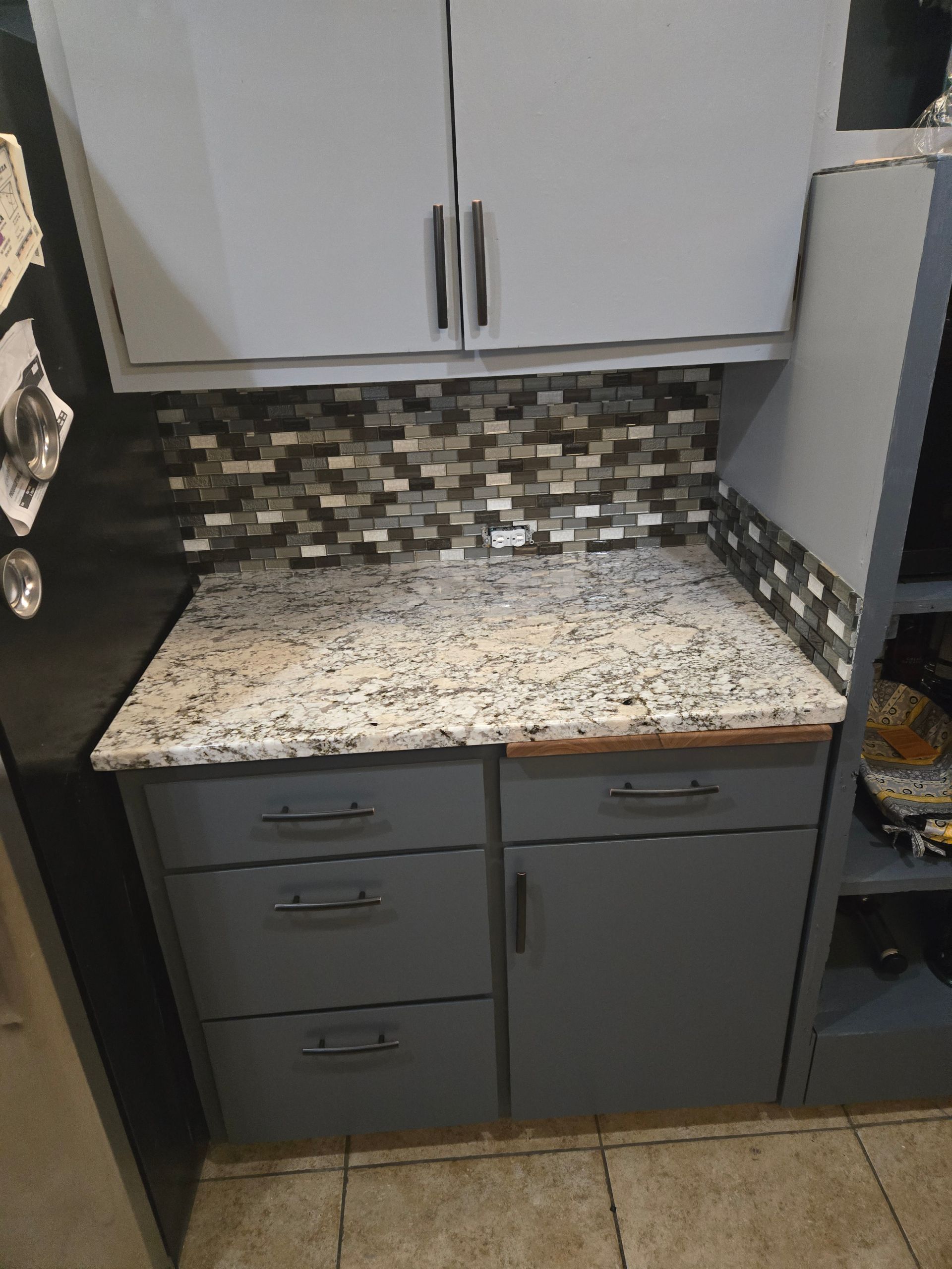 Kitchen counter with white-speckled top, mosaic backsplash, gray cabinets, and open shelving.