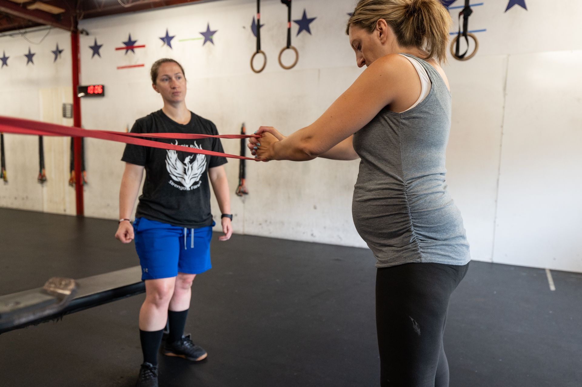 A pregnant woman is stretching her arms with a resistance band in a gym.