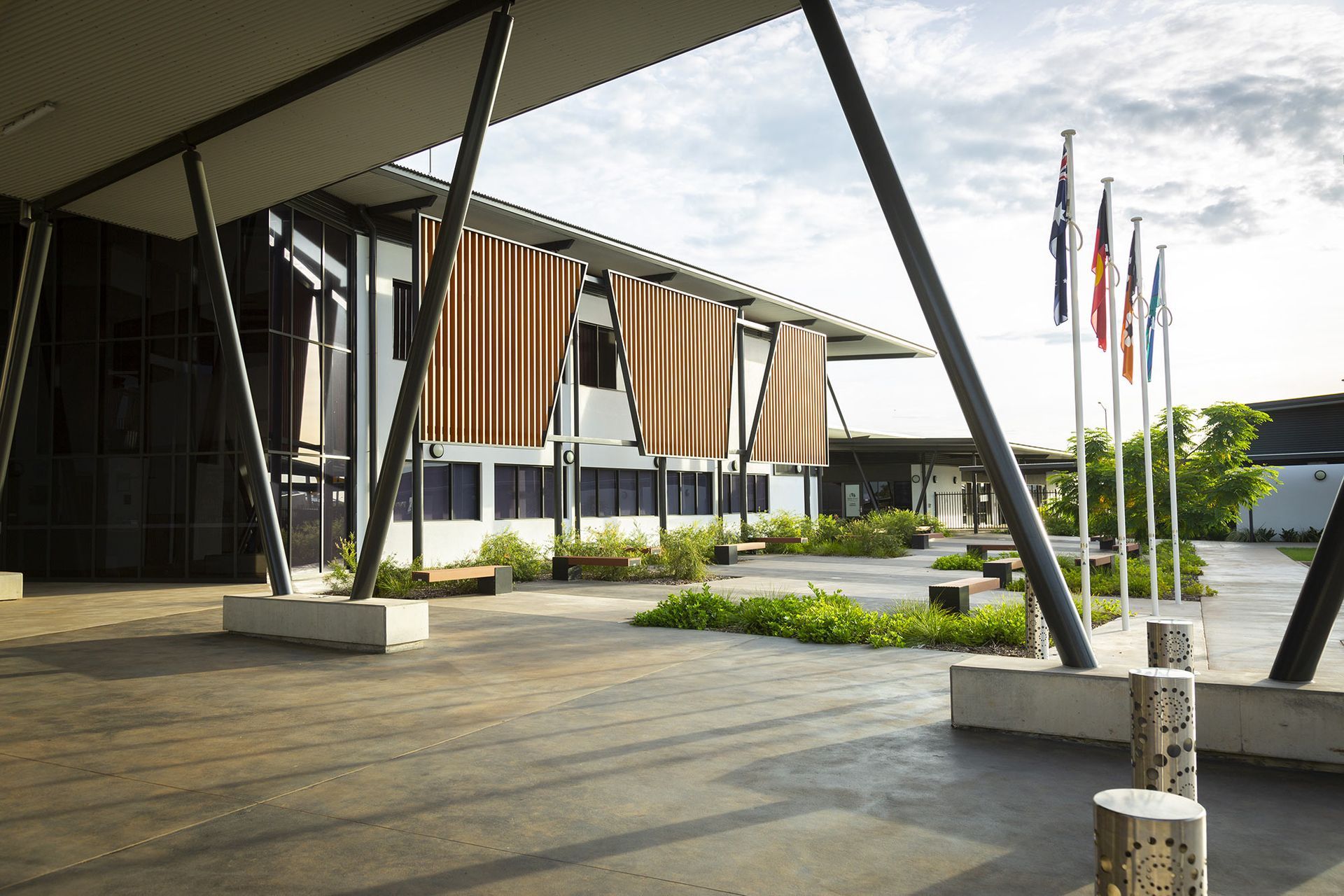 A modern building with large, bronze-colored hanging shades above a paved courtyard with flags and landscaping.