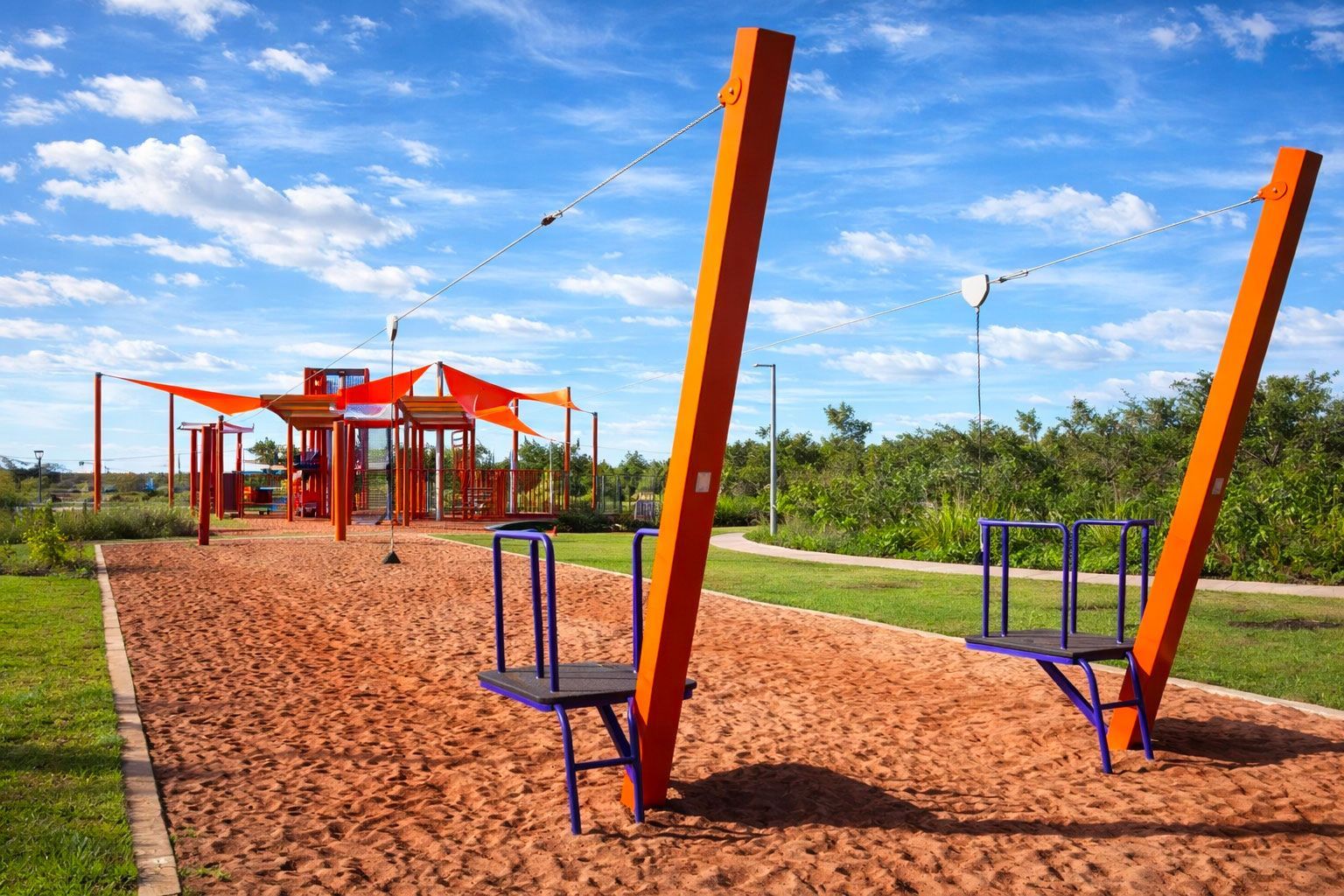 A playground featuring bright orange support beams, two metal platforms, and a shaded climbing structure on sandy ground.