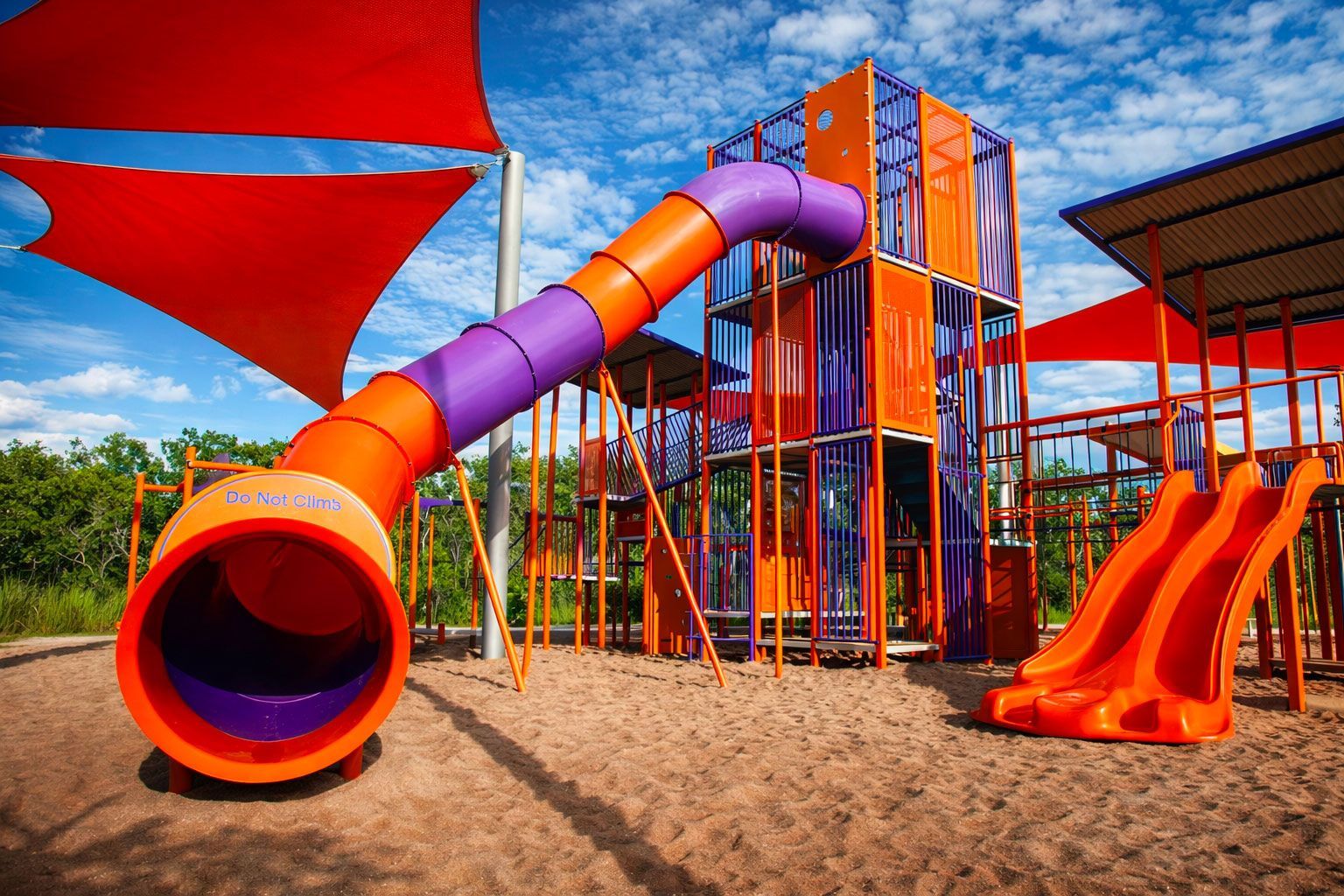 Colorful outdoor playground structure with a long tunnel slide and two side-by-side slides on a sandy ground.